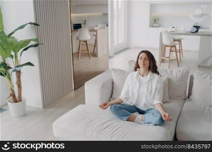 Young hispanic woman is practicing yoga and meditation at home. Girl in white is sitting on couch with her eyes closed. Posture exercise, lotus asana, gymnastics. Wellbeing and morning routine.. Young hispanic woman is practicing yoga and meditation at home. Wellbeing and morning routine.