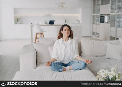 Young hispanic woman is practicing yoga and meditation on couch at home. Posture exercise, lotus asana. Morning gymnastics, healthy lifestyle. Concentration and zen finding concept.. Young hispanic woman in practicing yoga and meditation on couch at home. Zen finding.