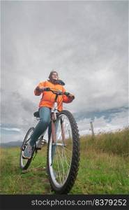 Young Hispanic woman seen from below wearing orange jacket and blue pants riding her bicycle alone through the rural countryside during a cloudy day. Young Hispanic woman wearing orange jacket and blue pants riding her bike through rural countryside