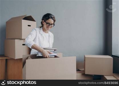 Young hispanic woman unpacking carton boxes and takes books out. Female student moves to c&us room. Happy girl in glasses unloading luggage. Relocation and entering to university.. Young hispanic woman unpacking carton boxes and takes books out. Entering to university.