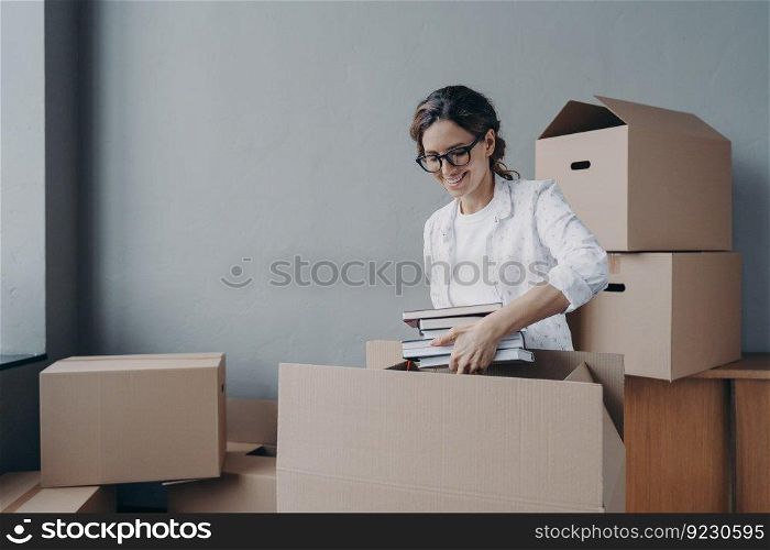 Young hispanic woman unpacking carton boxes and takes books out. Female student moves to campus room. Happy girl in glasses unloading luggage. Relocation and entering to university.. Young hispanic woman unpacking carton boxes and takes books out. Entering to university.