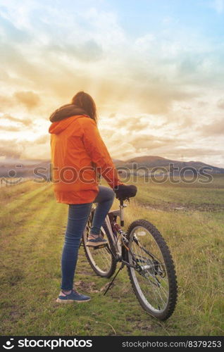 Young Hispanic woman wearing orange jacket and blue pants resting leaning on her bicycle seen from behind in the middle of the rural field during a sunset. Young Hispanic woman wearing an orange jacket and blue pants resting leaning on her bicycle in the middle of a rural field during a sunset