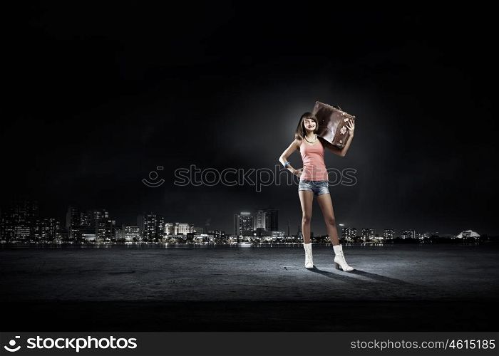 Young hitchhiking traveler. Young pretty girl with her retro suitcase on shoulder at night
