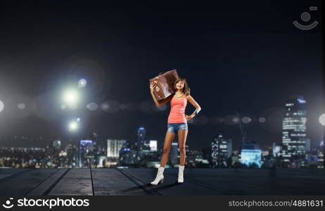 Young hitchhiking traveler. Young pretty girl with her retro suitcase on shoulder at night