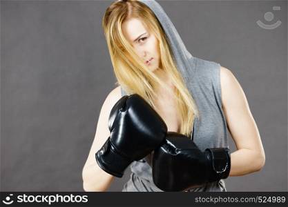 Young hooded woman fighting boxing. Blonde attractive girl wearing black punch gloves. Sport and fitness, power, exercising, on grey. Boxer girl exercise with boxing gloves.