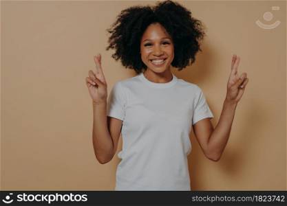Young hopeful african american female isolated on beige studio background crossing fingers and making wish, showing hope gesture and smiling at camera, asks for good luck. Superstition concept. Hopeful african american female isolated on beige studio background crossing fingers making wish