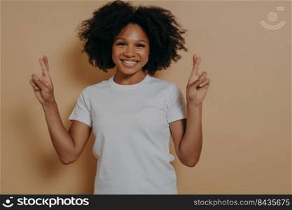 Young hopeful african american female isolated on beige studio background crossing fingers and making wish, showing hope gesture and smiling at camera, asks for good luck. Superstition concept. Hopeful african american female isolated on beige studio background crossing fingers making wish