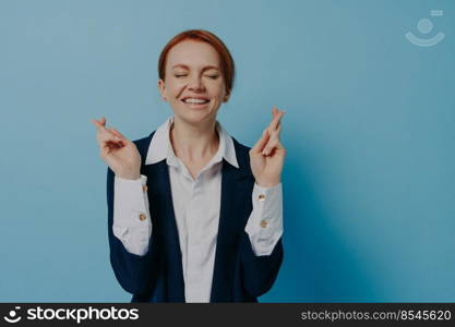Young hopeful red haired business woman with closed eyes in formal wear crossing fingers, wishing for luck, hoping for good result or something good happen, posing isolated over blue background. Young hopeful businesswoman with closed eyes in formal wear crossing fingers, isolated on blue