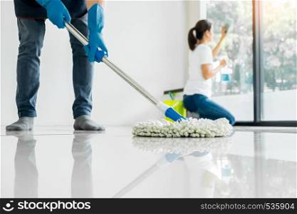 Young housekeeper cleaning floor mobbing holding mop and plastic bucket with brushes, gloves and detergents in the leaving room house floor helping his wife