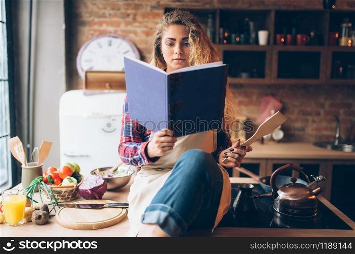 Young housewife in an apron reads recipe book, kitchen interior on background. Female cook prepares for cooking healthy vegetarian food, salad preparation. Young housewife in an apron reads recipe book