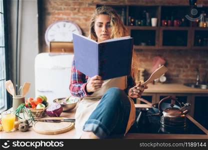 Young housewife in an apron reads recipe book, kitchen interior on background. Female cook prepares for cooking healthy vegetarian food, salad preparation. Young housewife in an apron reads recipe book