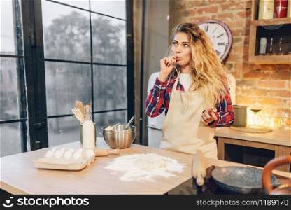 Young housewife in an apron stands against kitchen table with flour, eggs and milk. Female cook prepares fresh homemade cake. Domestic pie preparation. Housewife against table with flour, eggs and milk