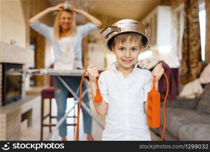 Young housewife irons the clothes, little boy dabbles with electricity. Woman doing housework at home. Female person with her playful son in the house