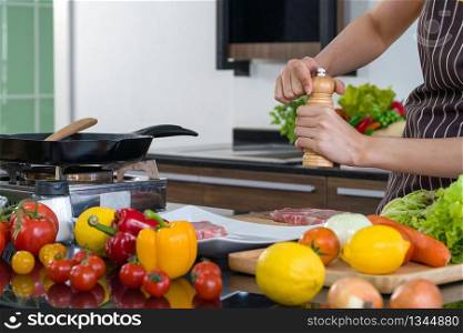 Young housewife putting pepper on the pork before cooking it in to a steak. Morning atmosphere in a modern kitchen. The kitchen counter full of various kinds of vegetables.