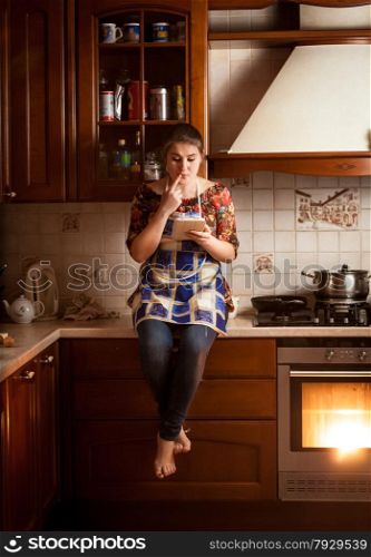 Young housewife sitting on tabletop and looking for recipe in tablet