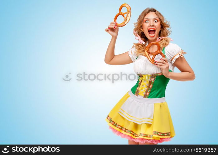 Young hungry sexy Oktoberfest woman wearing a traditional Bavarian dress dirndl holding two pretzels on blue background.