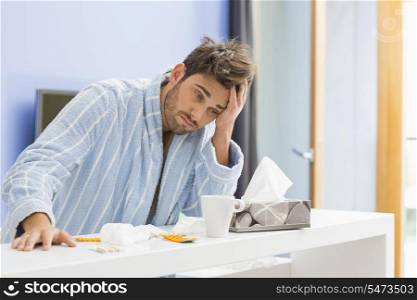 Young ill man with coffee mug; medicine and tissue leaning on kitchen counter