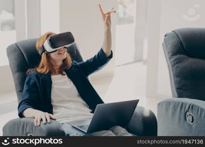 Young impressed redhead businesswoman using virtual reality glasses for business, female in VR headset pointing with finger in air, touching 3d object while working remotely on laptop from home. Female in VR headset pointing with finger, touching 3d object while working remotely on laptop