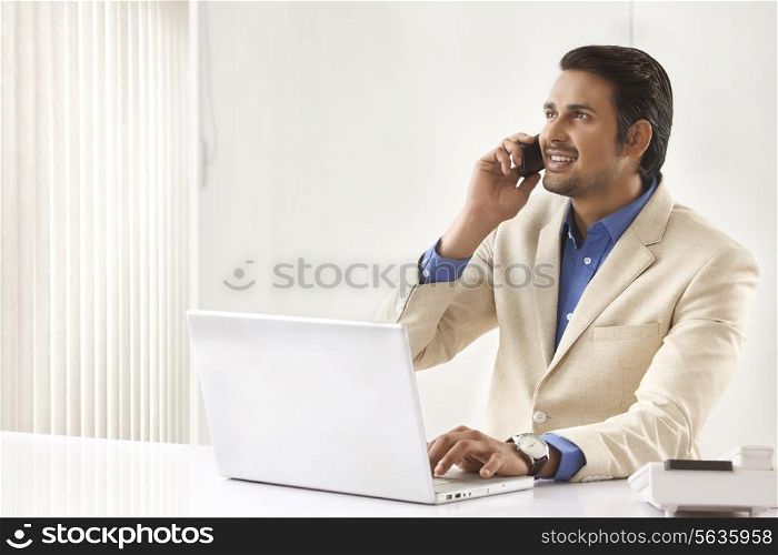 Young Indian businessman on call while using laptop in office