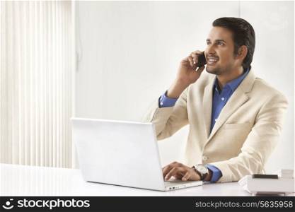Young Indian businessman on call while using laptop in office