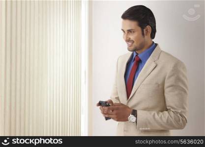 Young Indian businessman text messaging while looking through window blinds