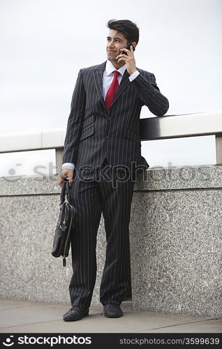 Young Indian businessman using cell phone with laptop bag by parapet