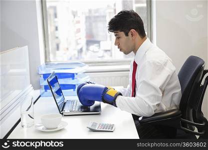 Young Indian businessman wearing boxing gloves while using laptop at office desk