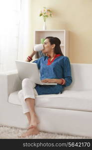 Young Indian woman with laptop drinking coffee on sofa