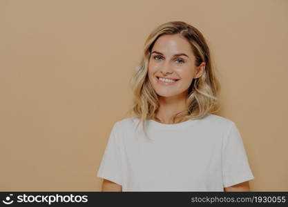 Young joyful woman with wavy blonde hair dressed casually feeling happy and smiling at camera, expressing happiness while standing isolated against beige studio wall. Face expressions concept. Young joyful woman with wavy blonde hair dressed casually feeling happy and smiling at camera, expressing happiness