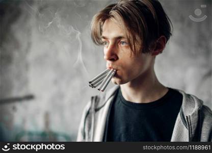 Young junkie with many cigarettes in his mouth reaching his hand forward, grunge background. No smoking concept, drug addict. Young junkie with many cigarettes in his mouth