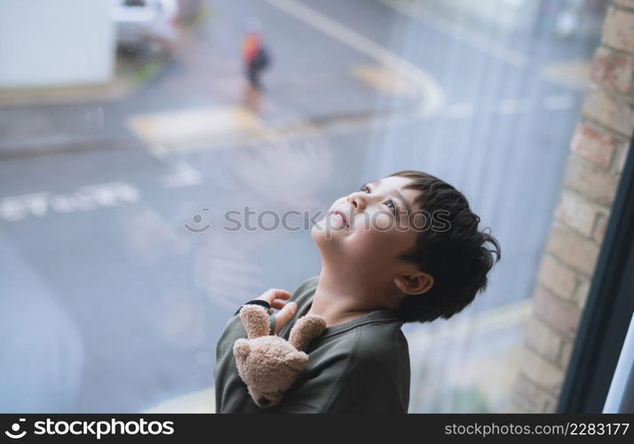 Young kid standing next to window looking up to sky with smiling face, Child looking through window glass with wondering face. Portrait boy relaxing in room apartment in city