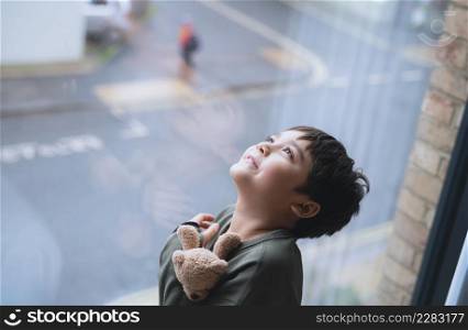 Young kid standing next to window looking up to sky with smiling face, Child looking through window glass with wondering face. Portrait boy relaxing in room apartment in city