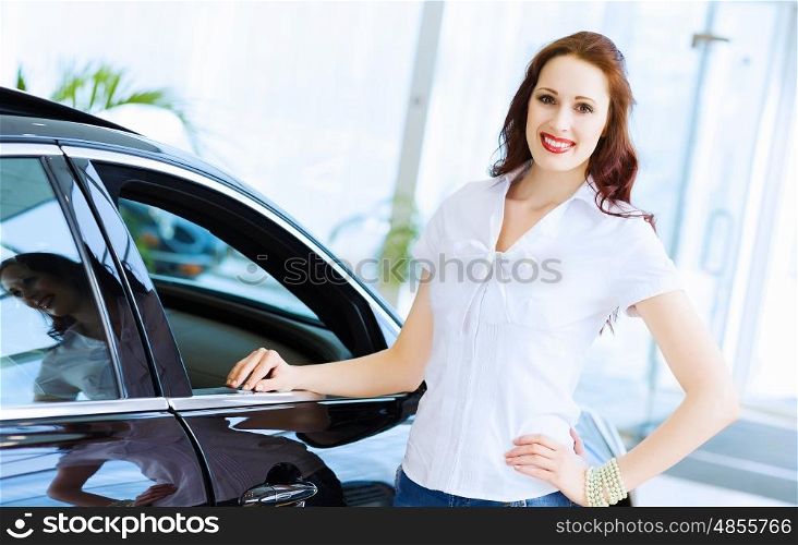 Young lady at car salon. Pretty woman standing near car at car center. Choosing a car