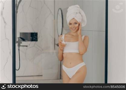 Young lady enjoying morning anti age anti wrinkle routine in bathroom after showering, smiling pretty woman wearing towel on head applying moisturizer cream under eye area. Beauty and skincare concept. Young lady enjoying morning anti age anti wrinkle routine in bathroom
