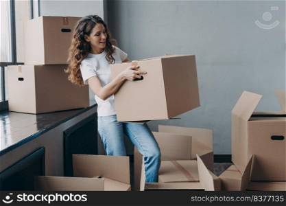 Young lady in white t-shirt is carrying cardboard box. Smiling attractive hispanic woman packing things and leaving. Packed boxes on the floor. Relocation and moving to new apartment concept.. Young lady in white t-shirt is carrying cardboard box. Hispanic woman packing things and leaving.