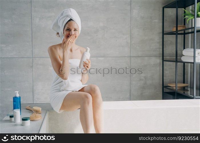 Young lady is relaxing in bathroom and laughing. Attractive european woman wrapped in towel applying body lotion after bathing. Girl takes shower at home. Anti-cellulite massage and spa.. Young lady is relaxing in bathroom and laughing. Woman wrapped in towel applying body lotion.