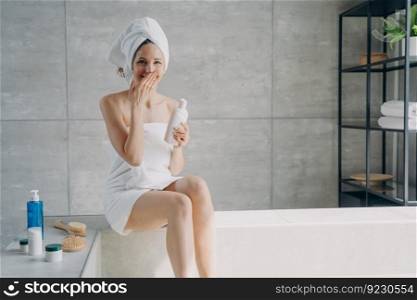 Young lady is relaxing in bathroom and laughing. Attractive european woman wrapped in towel applying body lotion after bathing. Girl takes shower at home. Anti-cellulite massage and spa.. Young lady is relaxing in bathroom and laughing. Woman wrapped in towel applying body lotion.