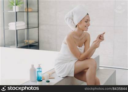 Young lady relaxing at spa resort. Girl reading cosmetics ingredients list on the pack and smiling. Caucasian woman wrapped in towel sitting on bathtub. Concept of natural cosmetics, skin treatment.. Young lady relaxing at spa resort. Girl reading cosmetics ingredients list on the pack and smiling.