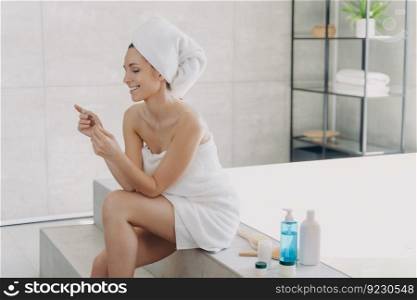 Young lady relaxing at spa resort. Girl reading cosmetics ingredients list on the pack and smiling. Caucasian woman wrapped in towel sitting on bathtub. Concept of natural cosmetics, skin treatment.. Young lady relaxing at spa resort. Girl reading cosmetics ingredients list on the pack and smiling.