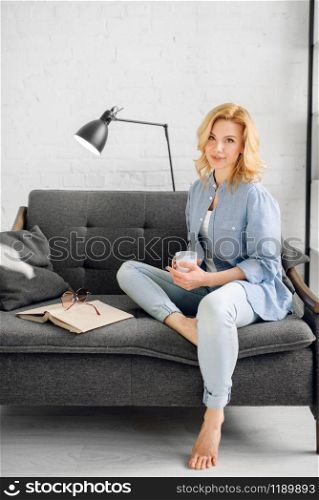 Young lady with book and cup of coffee poses on cozy black couch, living room in white tones on background. Attractive female person with magazine sitting on sofa at home. Lady with book and cup of coffee on black couch