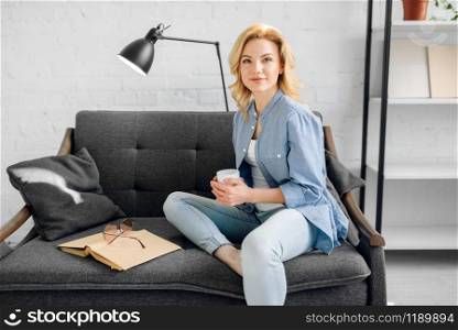 Young lady with book and cup of coffee poses on cozy black couch, living room in white tones on background. Attractive female person with magazine sitting on sofa at home