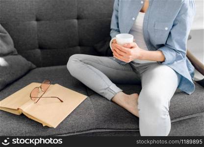 Young lady with book and cup of coffee poses on cozy black couch, living room in white tones on background. Attractive female person with magazine sitting on sofa at home. Lady with book and cup of coffee on black couch