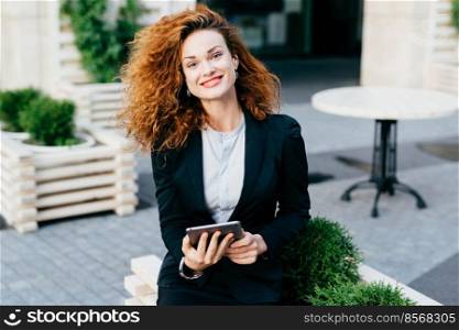 Young lady with curly hair, red painted lips and shining eyes, wearing black suit and shirt, sitting at outdoor cafe with tablet, smiling pleasantly into camera. Attractive female at terrace
