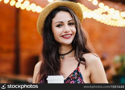 Young lady with make-up, wearing straw hat, necklace and dress, holding paper cup with coffee, smiling pleasantly at camera while posing at restaurant. Woman with pleased expression resting at cafe