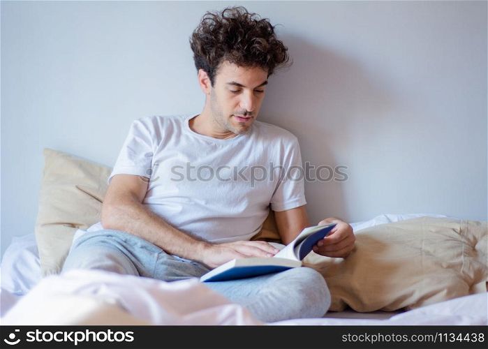 Young latin man reading a book while laying in bed at home.