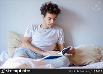 Young latin man reading a book while laying in bed at home.
