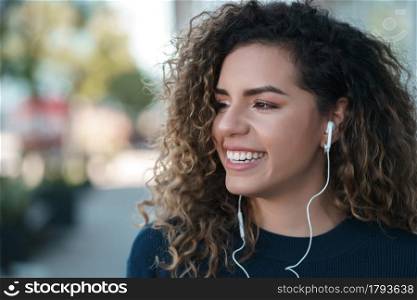 Young latin woman listening to music with earphones while standing outdoors on the street. Urban concept.