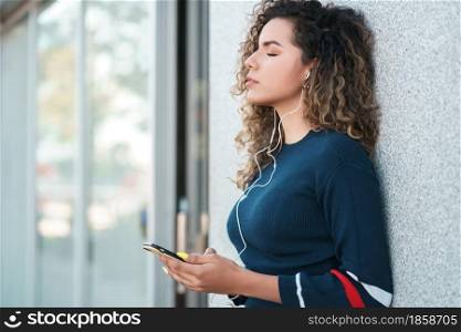 Young latin woman listening to music with her mobile phone while standing outdoors on the street. Urban concept.