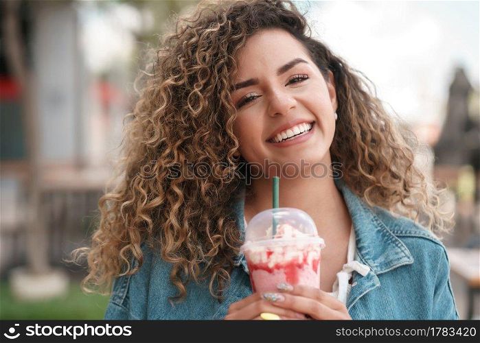 Young latin woman smiling and looking at the camera while drinking a cold drink at a coffee shop outdoors on the street. Urban concept.