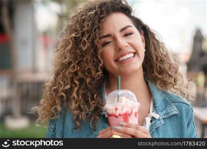 Young latin woman smiling and looking at the camera while drinking a cold drink at a coffee shop outdoors on the street. Urban concept.